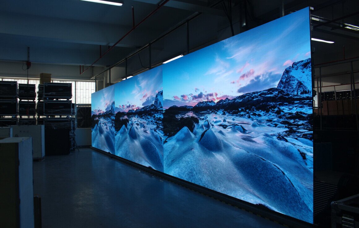 LED Wall Panoramico Un grande LED wall panoramico che mostra un paesaggio invernale mozzafiato con rocce innevate e un cielo al tramonto vibrante, installato in un ambiente industriale con scaffali e attrezzature sullo sfondo.